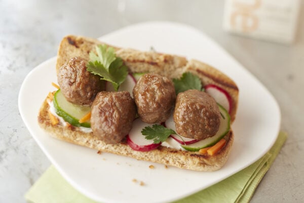 Sandwich containing meatballs, basil, radishes and carrots on a sub roll on top of a white plate with a light green napkin underneath.