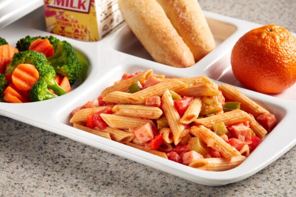 A white tray with penne pastalaya, an orange, breadsticks milk, and a mix of broccoli and carrots