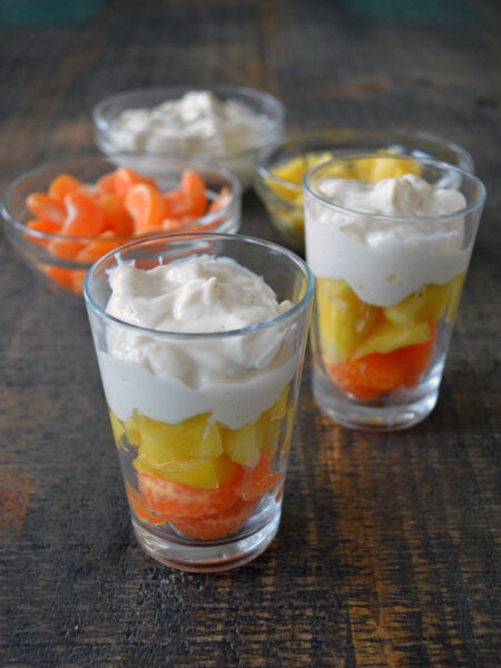 Two clear glasses with mandarin oranges, pineapple and Greek yogurt layered and three clear bowls, one with mandarin oranges, one with Greek yogurt and one with Pineapple on a brown, wood surface