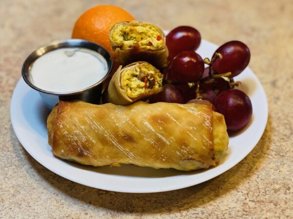Two egg rolls on a white plate with grapes, an orange, and a cup of white suace