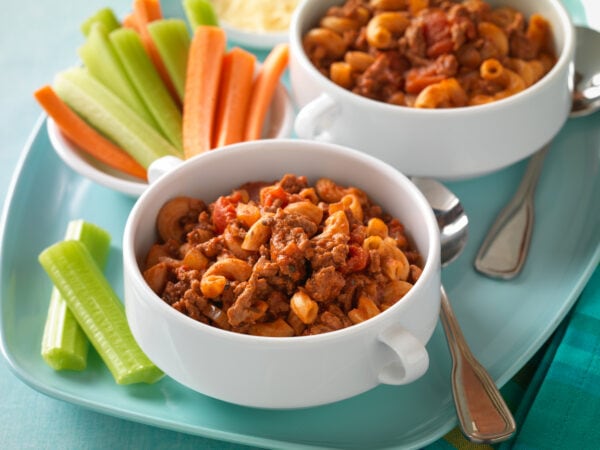 two bowls of goulash, a bowl of celery and carrots and two spoons on a blue plate