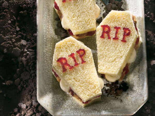 Three banana PB&J "casket" sandwiches with "RIP" written on top sitting on a grey plate sprinkled with crumbled cookies