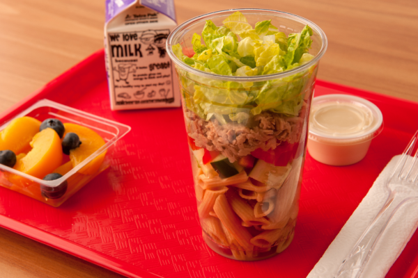 Clear cup with pasta, cucumber, red bell pepper, tuna and shredded lettuce on a red tray with sides of fruit, milk and salad dressing