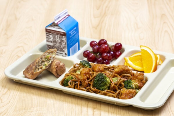 Noodles with chicken and broccoli on a white tray with an eggroll, grapes, orange slices and chocolate milk