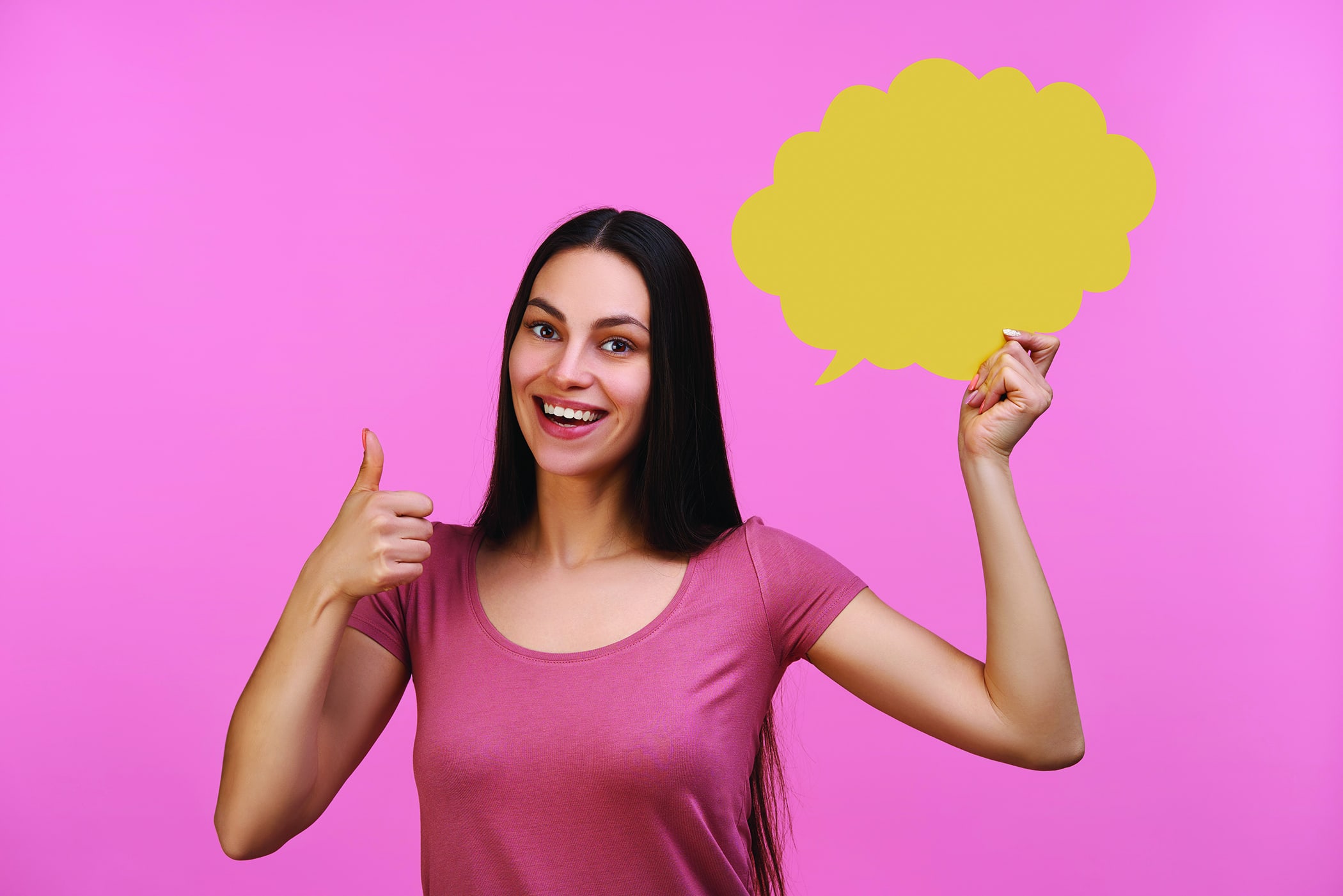 Young excited smiling girl student holding blank yellow speech cloud in hand and showing thumbs up gesture. Isolated over pastel pink background.