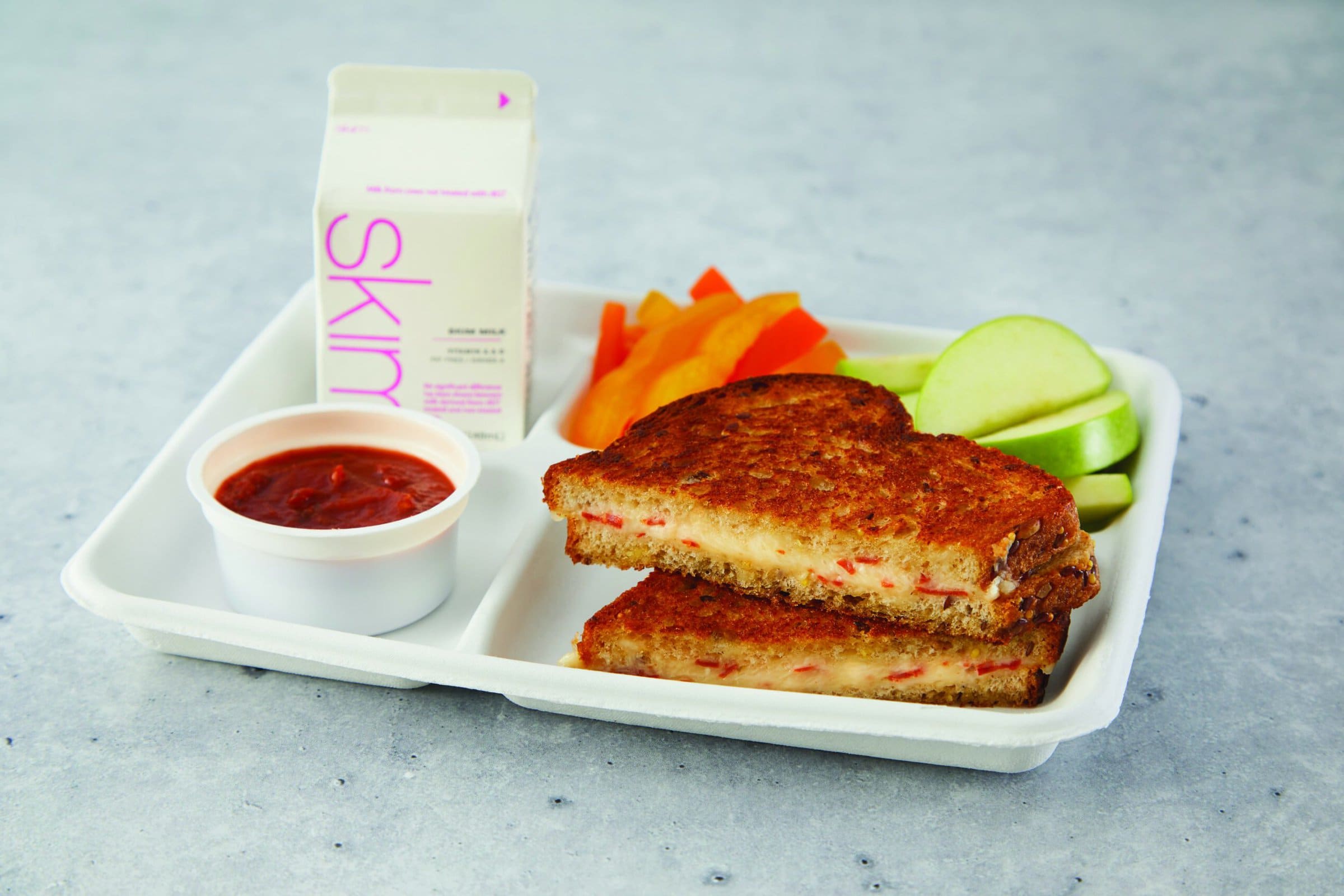 Grilled cheese and pepperoni sandwich on a white tray next to apple slices, a carton of skim milk and a cup of pizza sauce