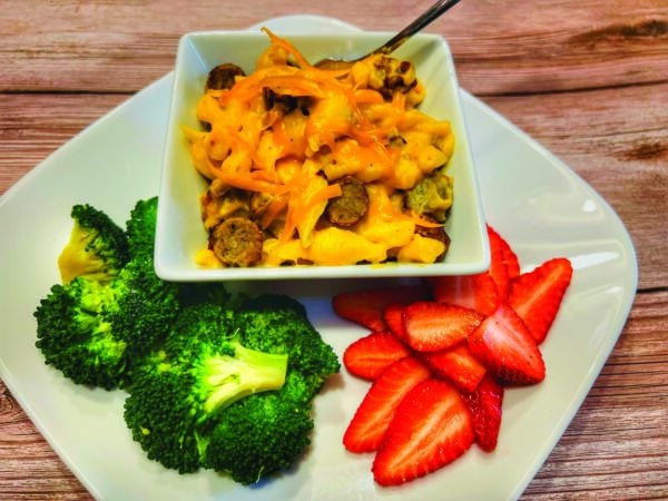 A bowl of sausage mac and cheese served on a plate with broccoli and strawberries