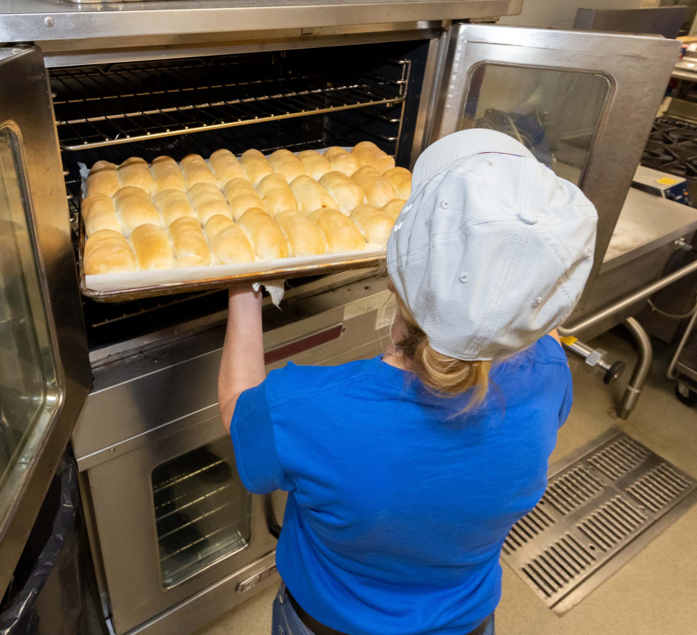 Woman in blue shirt takes out dinner rolls from a large oven.